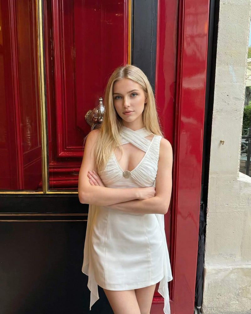 A blonde model with arms crossed posing in front of an elegant red door, wearing an eclipse white shell dress featuring a ruched bodice and draping details.