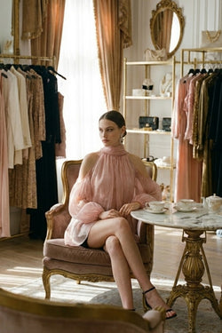 Woman in aurorette light pink tulle top sitting in an elegant room with clothing racks and a table.