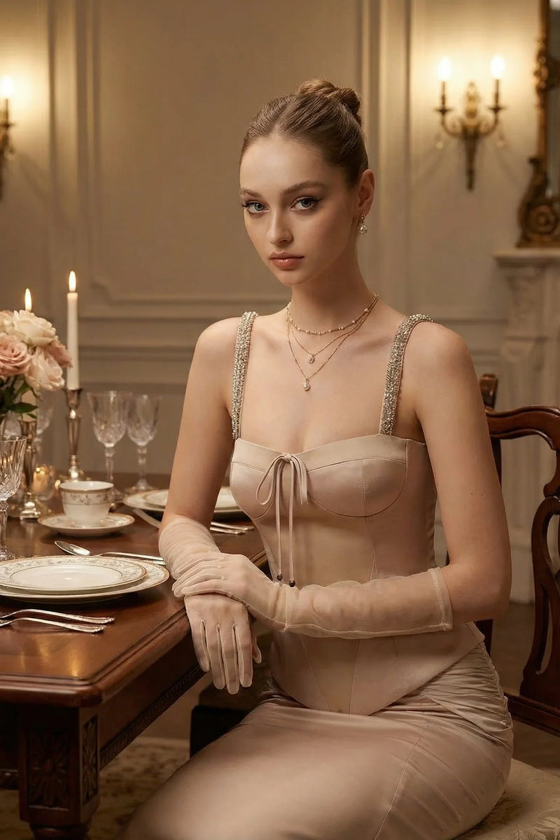 A model sitting at a candlelit dining table wearing sheer gloves, an embellished corset top, and a flowing satin maxi skirt, showcasing romantic evening elegance.